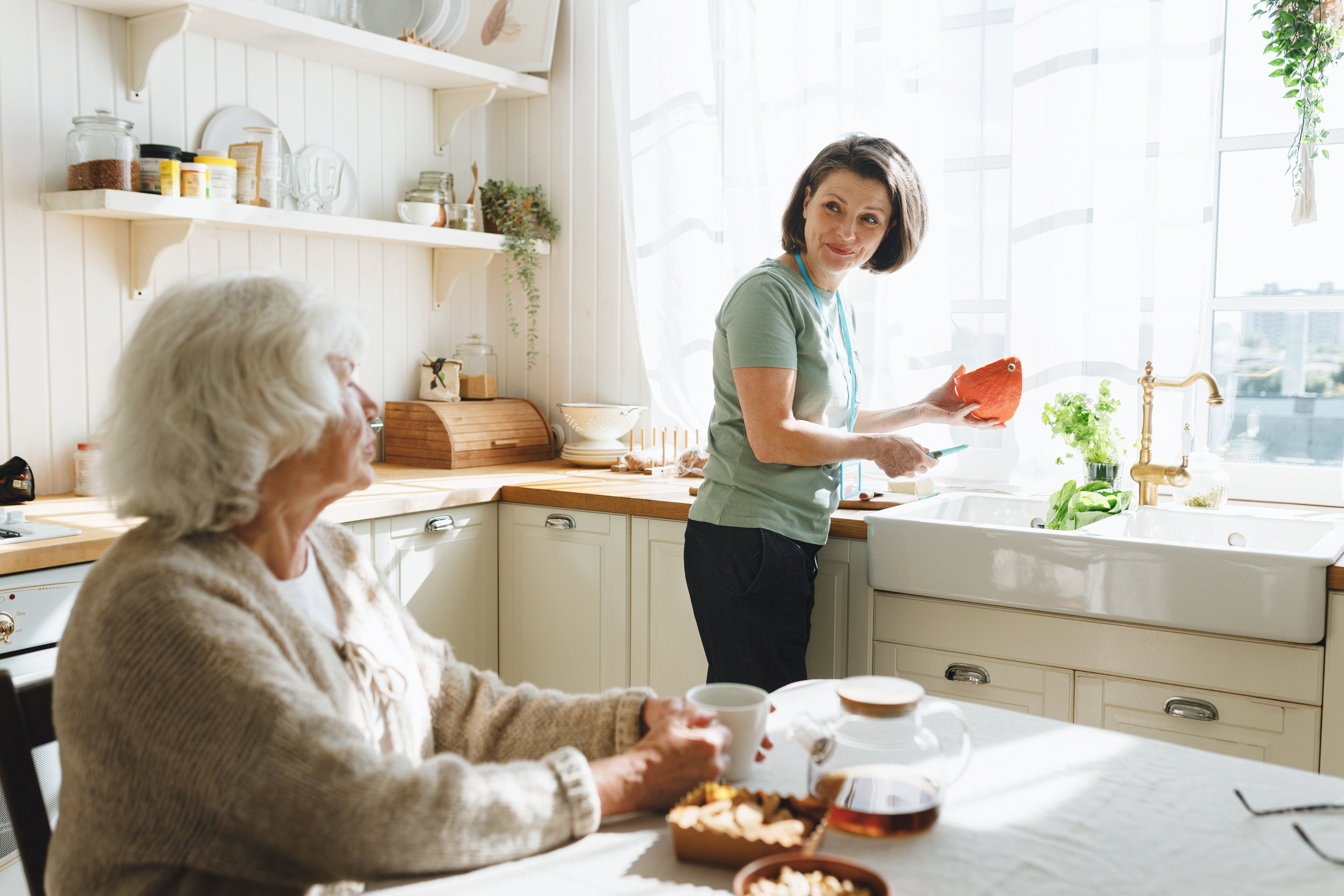 A caregiver and an older woman together in a home kitchen, sharing a quiet moment during the day.